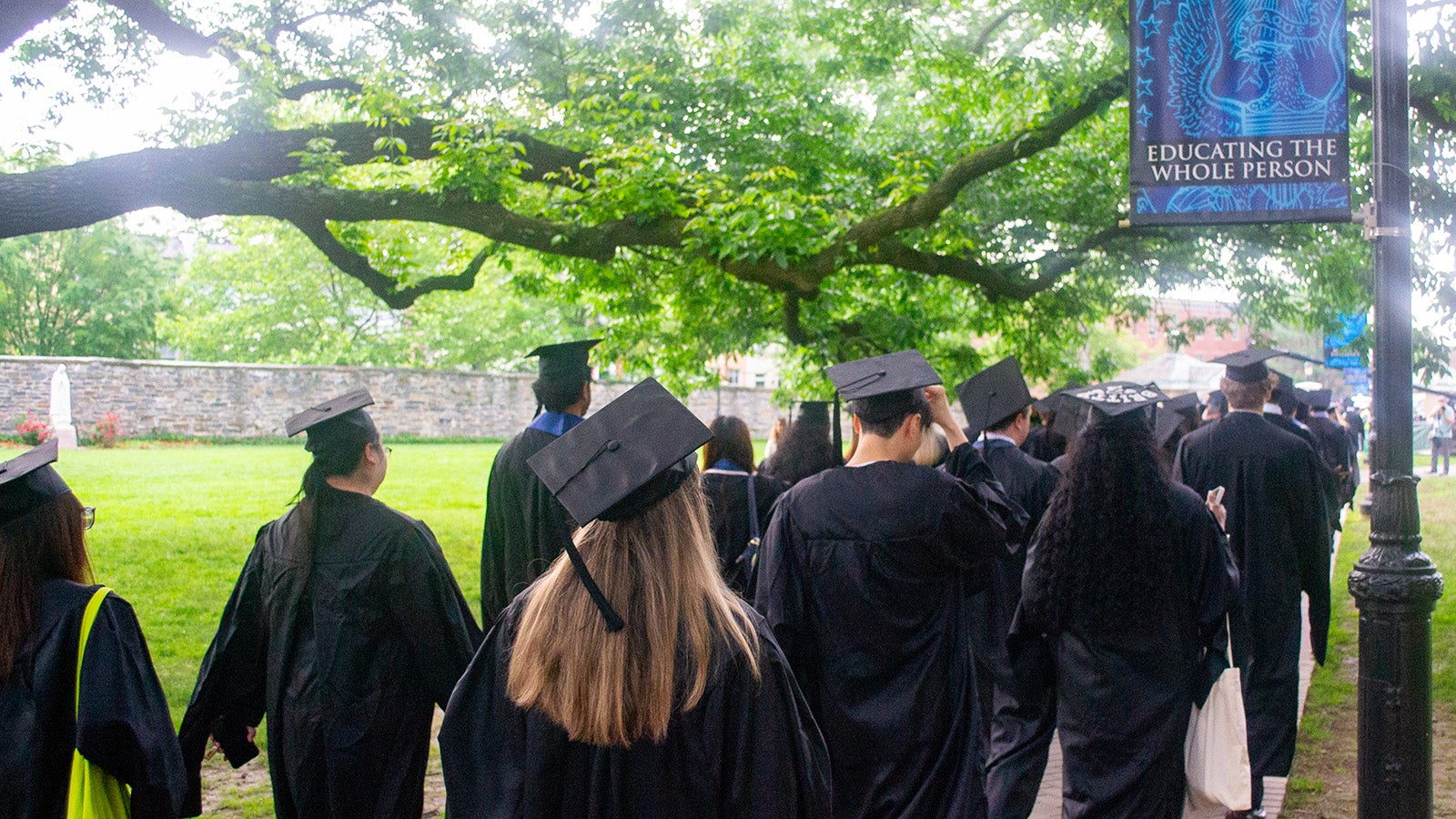 Graduates wearing their regalia process toward the Commencement tent under a Georgetown banner with the words "Educating the Whole Person"